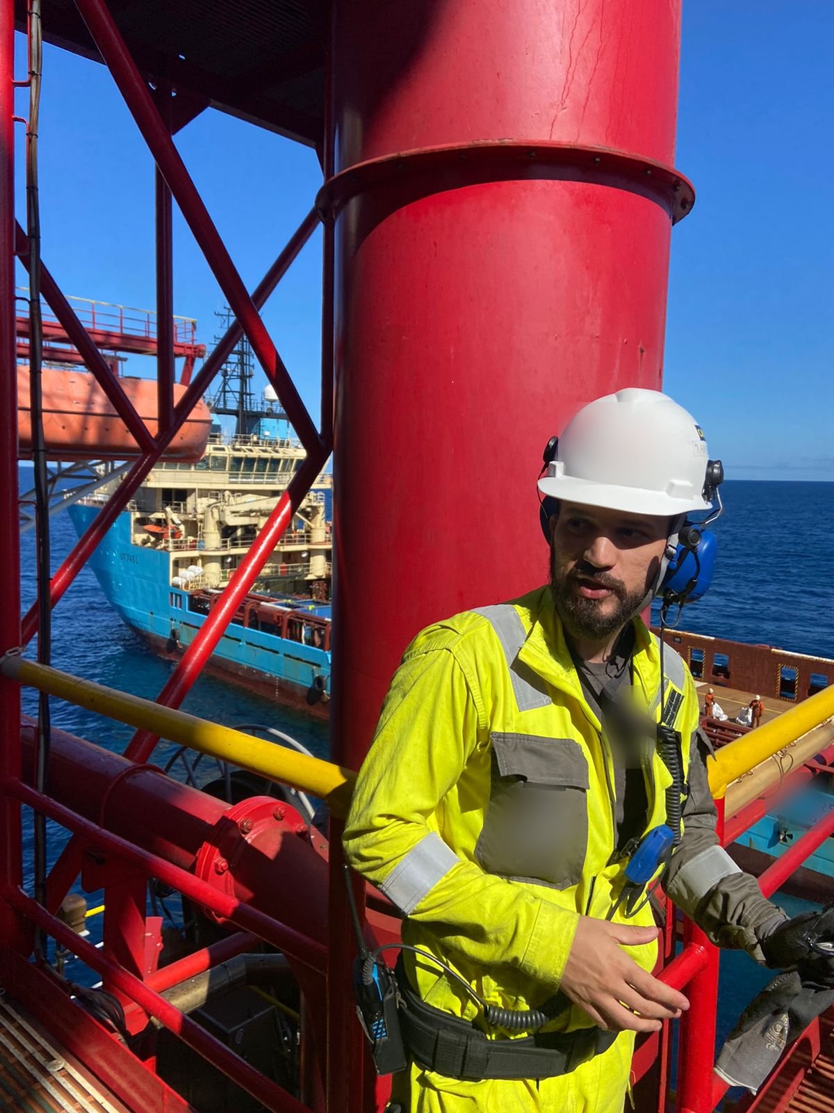 Rafael de macacão e capacete no convés, com embarcação de apoio ao fundo